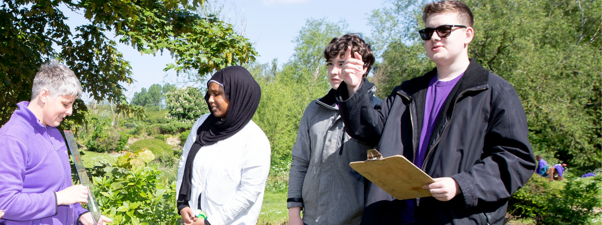 A group stood outside, a teacher giving an explanation to 3 students, one with a clipboard