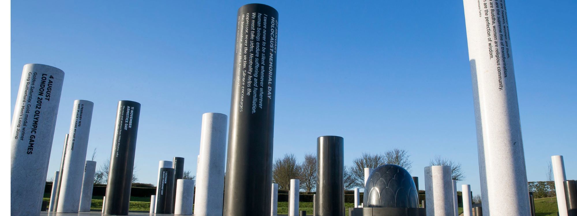 The image depicts an outdoor memorial featuring cylindrical pillars of varying heights arranged on a tiled platform under a clear blue sky. Each pillar is inscribed with important dates or events, such as "HOLOCAUST MEMORIAL DAY" and "LONDON 2012 OLYMPIC GAMES." The installation includes a dome-like structure in the background, with trees and greenery visible. The design invites reflection on significant historical moments.