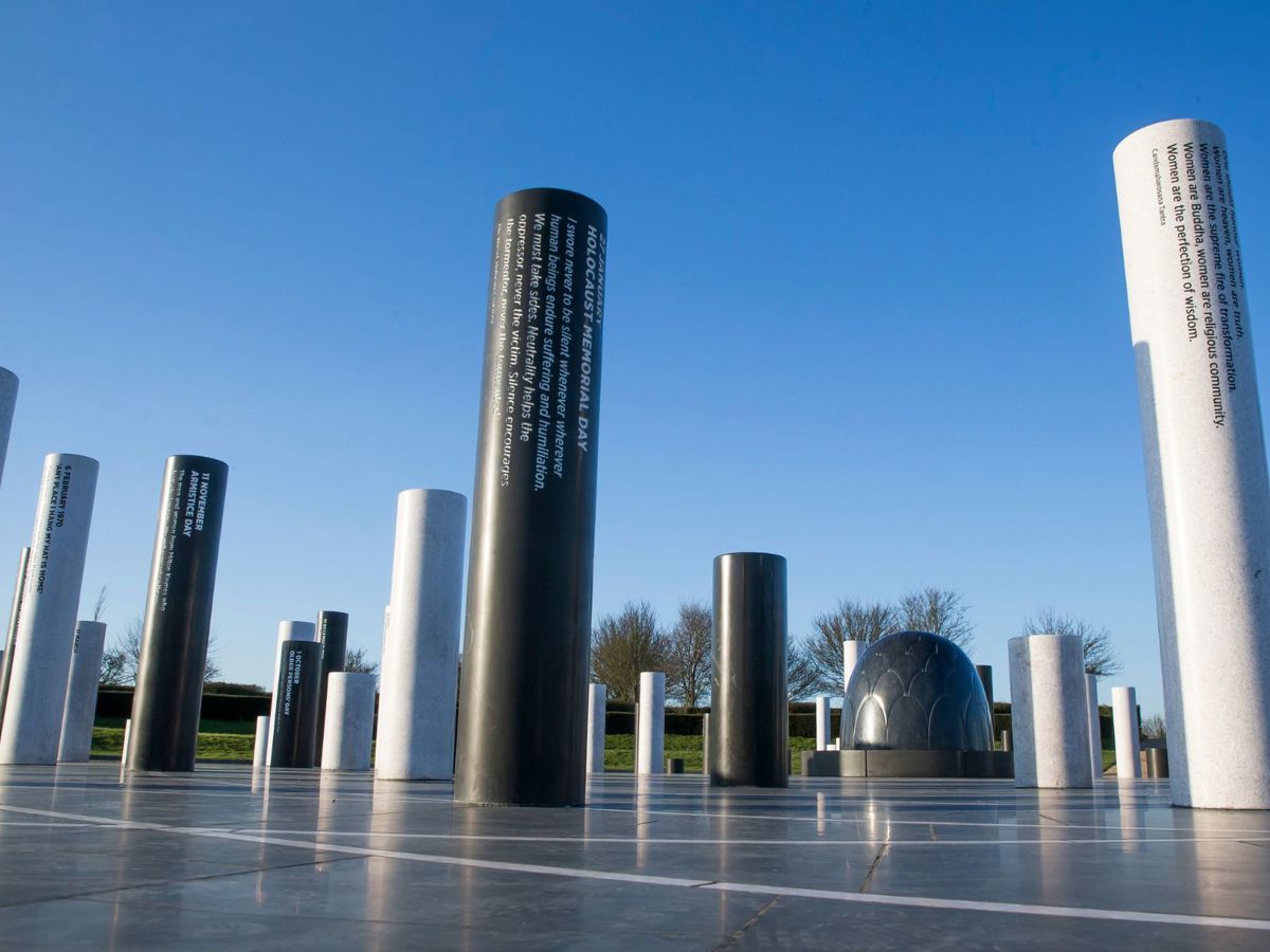 The image depicts an outdoor memorial featuring cylindrical pillars of varying heights arranged on a tiled platform under a clear blue sky. Each pillar is inscribed with important dates or events, such as "HOLOCAUST MEMORIAL DAY" and "LONDON 2012 OLYMPIC GAMES." The installation includes a dome-like structure in the background, with trees and greenery visible. The design invites reflection on significant historical moments.