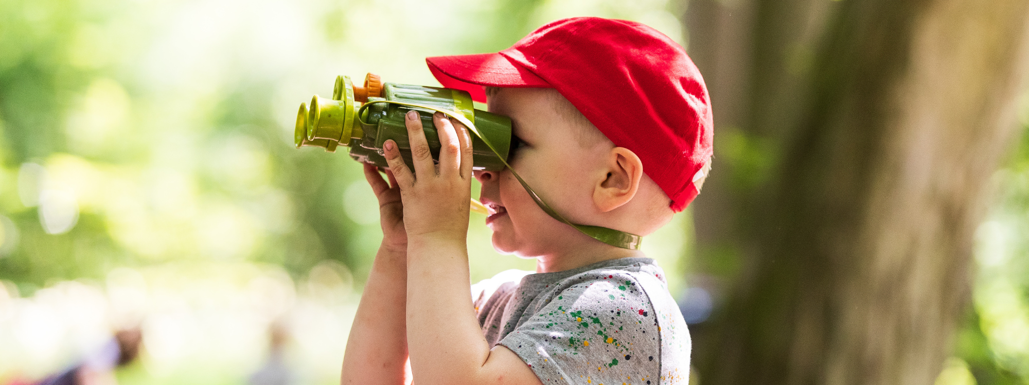 A young boy wearing a red summer hat using some binoculars in a sunny woodland