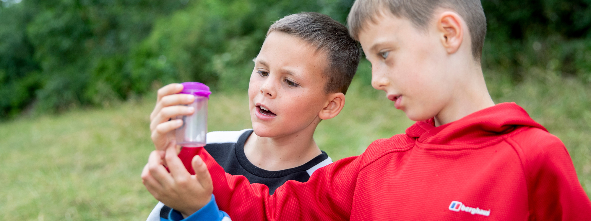 Two young boys stare inquisitively into a small plastic container which has a small insect inside 