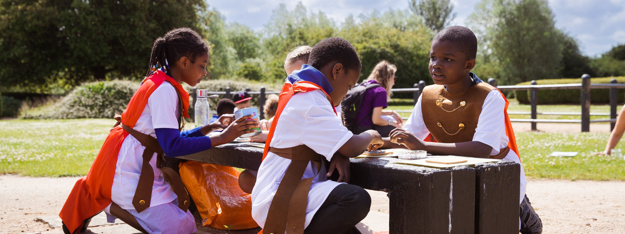 Children wearing Roman costumes engage in an activity at a bench in the sunshine