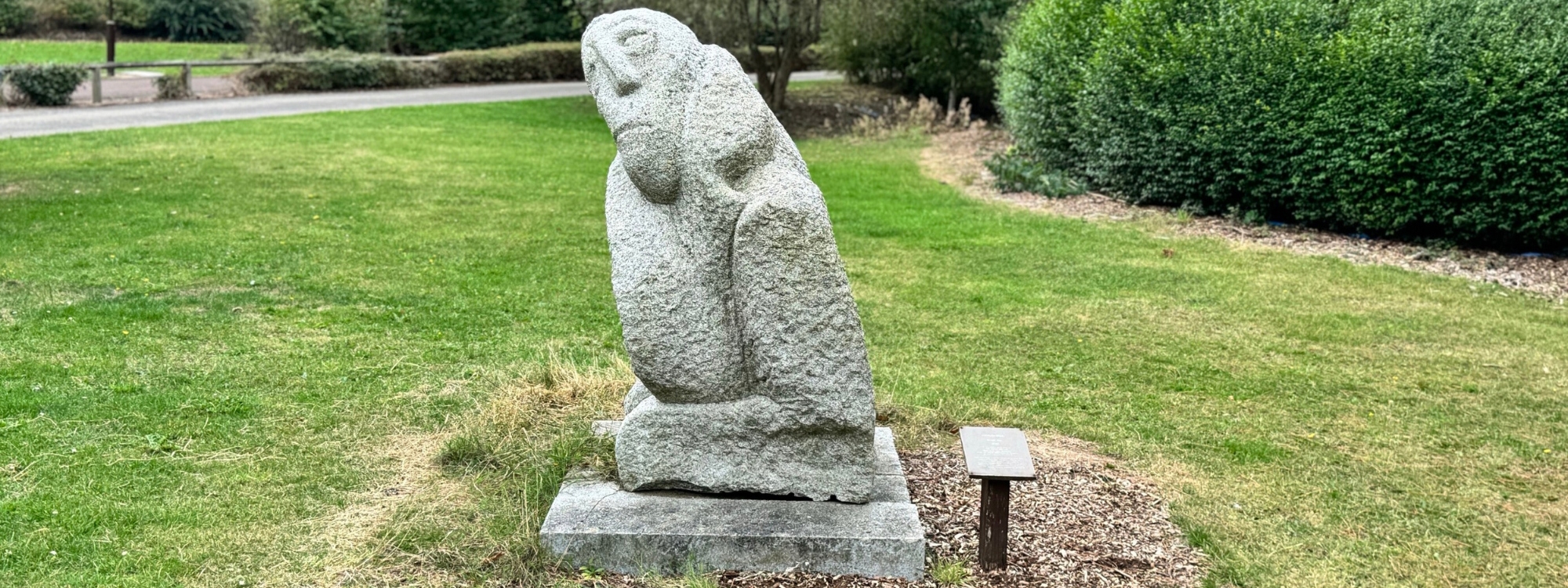 The image shows a stone sculpture in a park or garden setting. The sculpture appears abstract, with smooth, rounded contours resembling a seated or crouched figure. It is placed on a square stone base, and there is a small plaque or sign mounted on a short post nearby, likely providing details about the artwork. The surrounding area consists of green grass, trimmed bushes, and trees in the background, giving it a peaceful, natural ambiance.