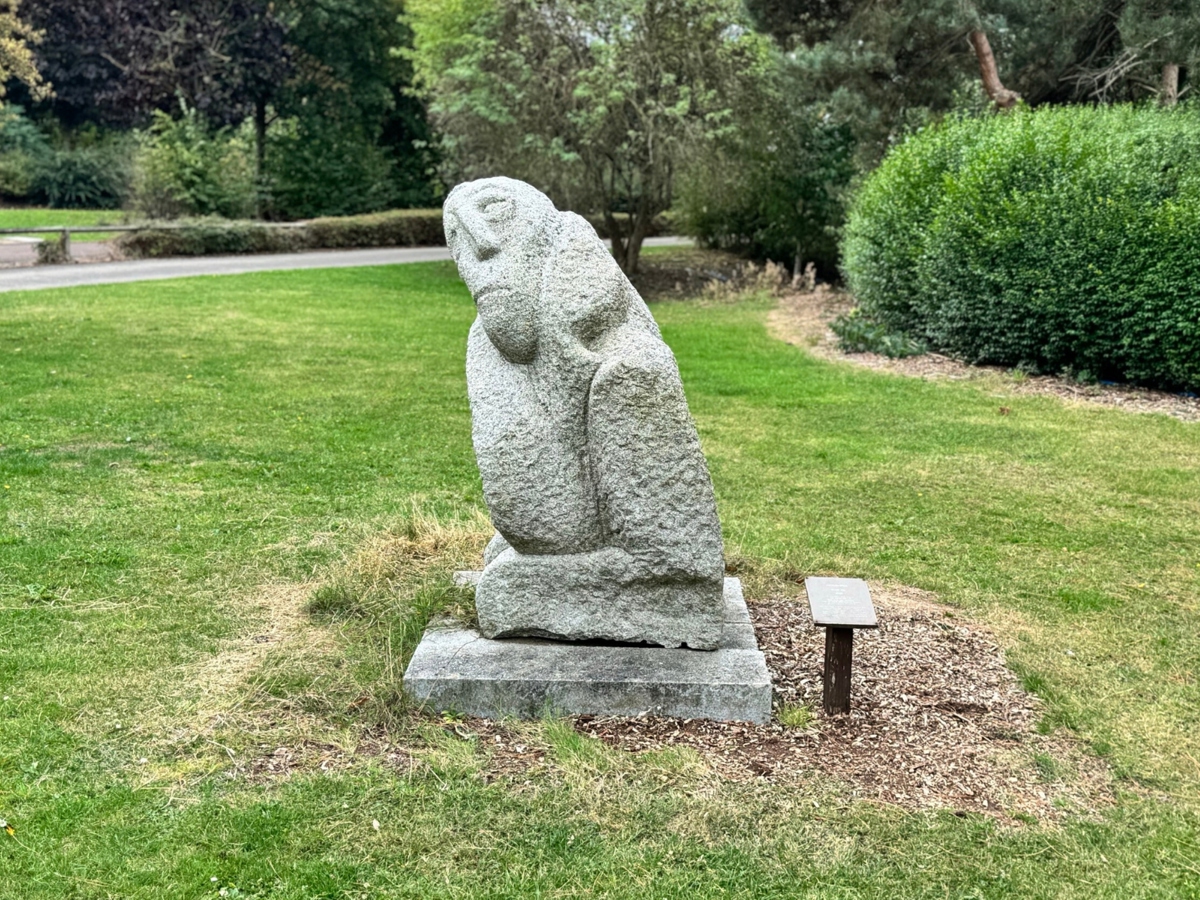 The image shows a stone sculpture in a park or garden setting. The sculpture appears abstract, with smooth, rounded contours resembling a seated or crouched figure. It is placed on a square stone base, and there is a small plaque or sign mounted on a short post nearby, likely providing details about the artwork. The surrounding area consists of green grass, trimmed bushes, and trees in the background, giving it a peaceful, natural ambiance.