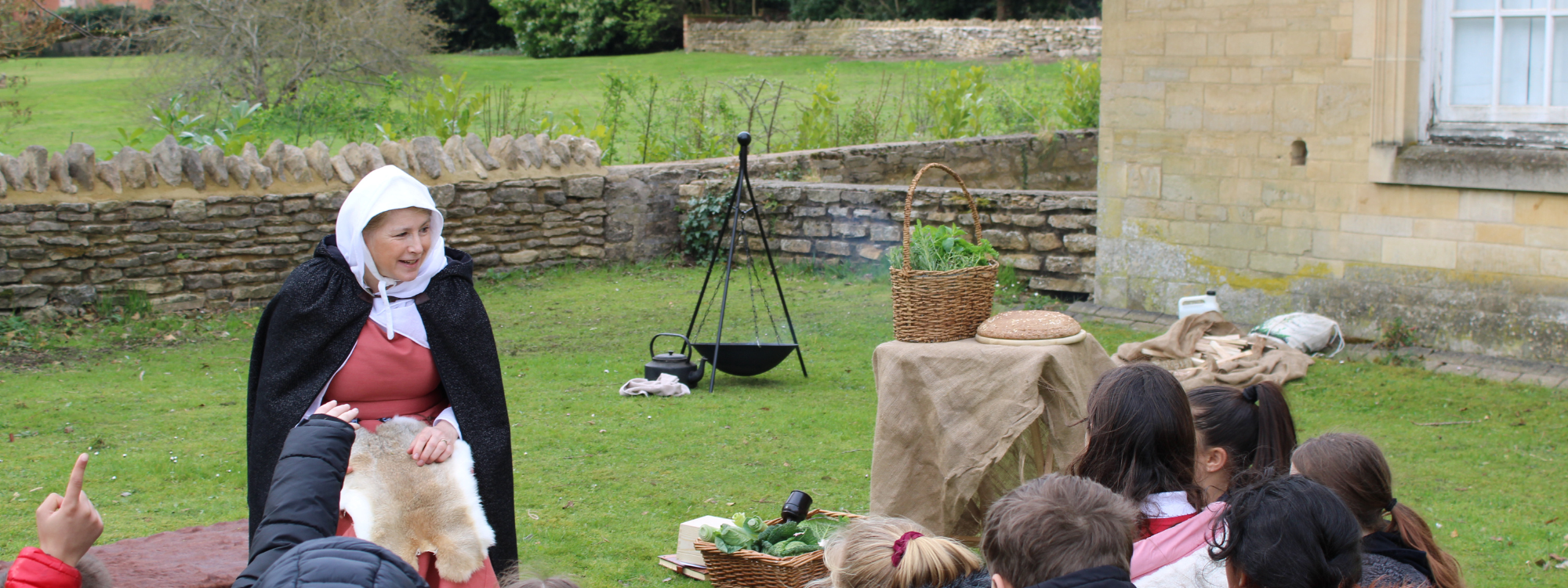 A lady in medieval costume is talking to a group of school students. They are sat on the grass out the front of an old building. 