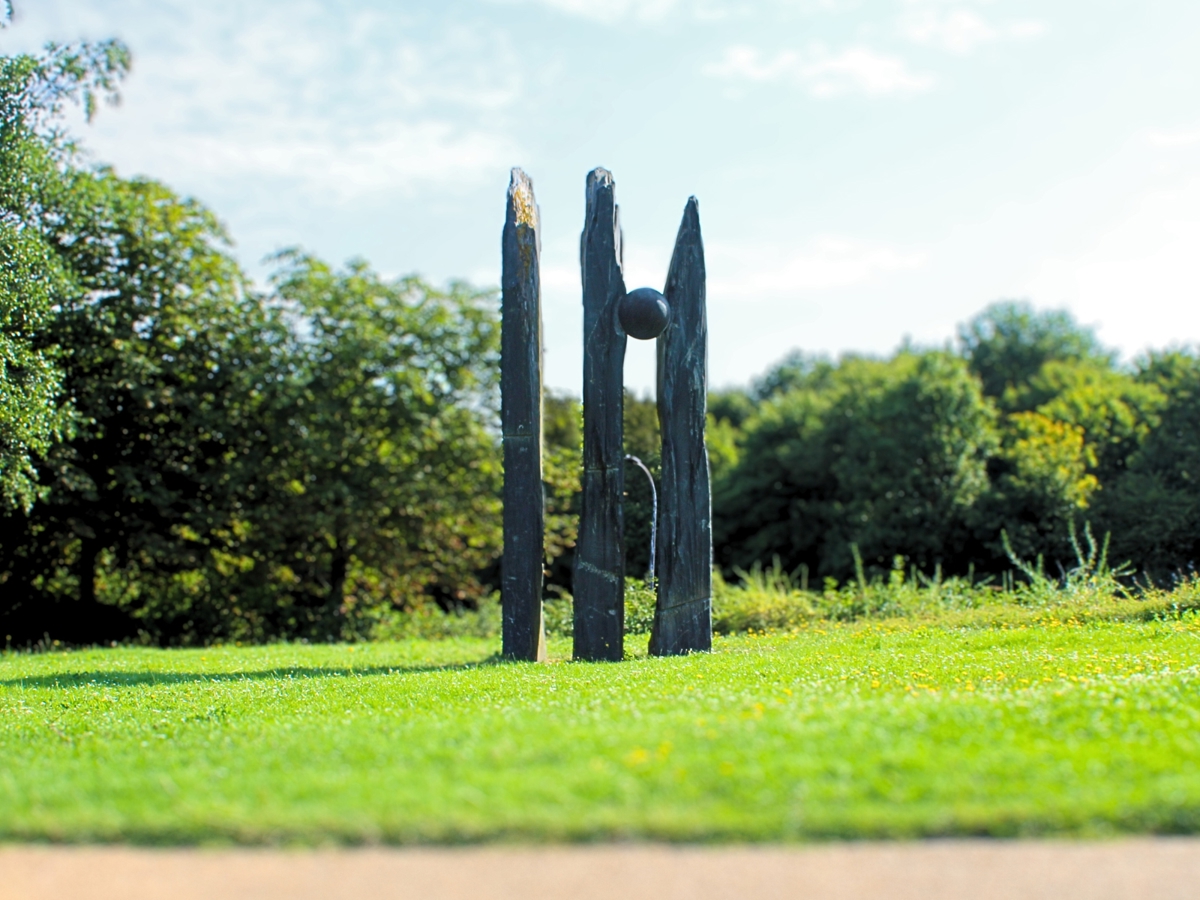 this stone structure has 3  varying vertical rectangular poles however between the 2nd a 3rd pole a large stone ball has been merged into the gap between the poles