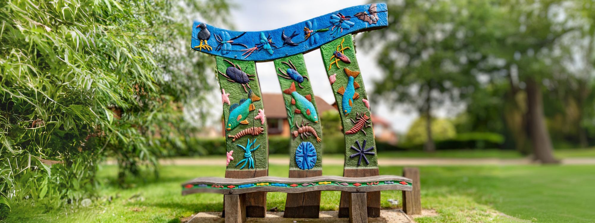 A colorful, artistic bench in a park, featuring a vibrant design with three tall, curved backrests adorned with intricate carvings of aquatic and insect life. The carvings include fish, birds, dragonflies, and other creatures in bright blue, green, and red hues. The bench seat itself is wavy, continuing the nature-inspired theme with additional small, colorful carvings. Surrounding the bench are lush green trees and a well-kept lawn, with a house visible in the background.