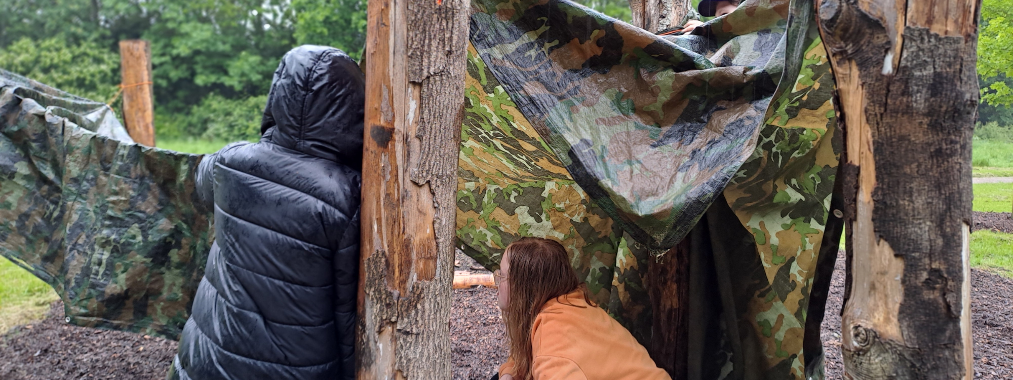 Children in the rain building shelters between tree trunks with camouflage tarpaulin