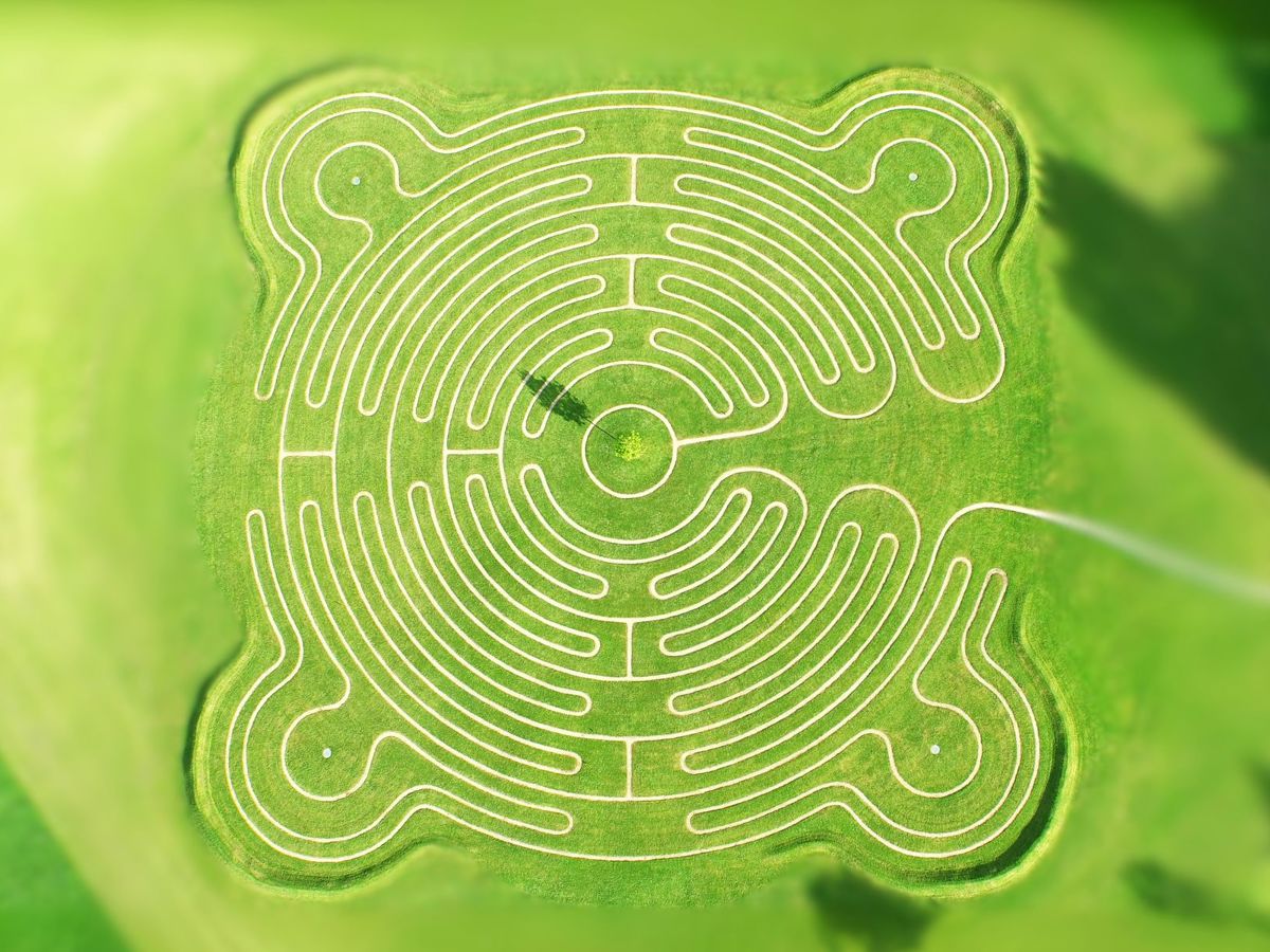 An aerial view of a large, circular labyrinth set in a grassy field. The labyrinth features intricate, symmetrical pathways outlined in white, forming a complex yet harmonious pattern. The design has four rounded corners that extend outward, adding to its uniqueness. At the center of the labyrinth is a small circular area with a tree, casting a shadow. The surrounding area is lush green, and the labyrinth is perfectly integrated into the landscape, offering a tranquil and meditative visual experience.