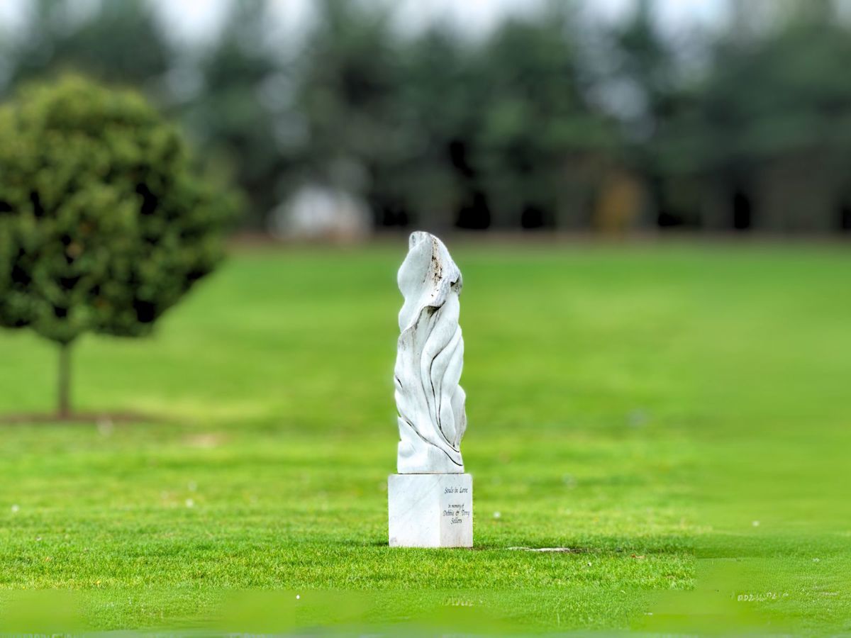 A serene park scene with a lush green lawn and a single, rounded tree on the left. On the right stands a smooth, white marble sculpture with abstract, flowing lines, possibly representing intertwined forms. The sculpture is inscribed with "Souls in Love. In memory of Debbie & Tony Salam." In the background, a row of tall, dark evergreen trees adds depth and contrast. The image exudes a peaceful and reflective atmosphere.