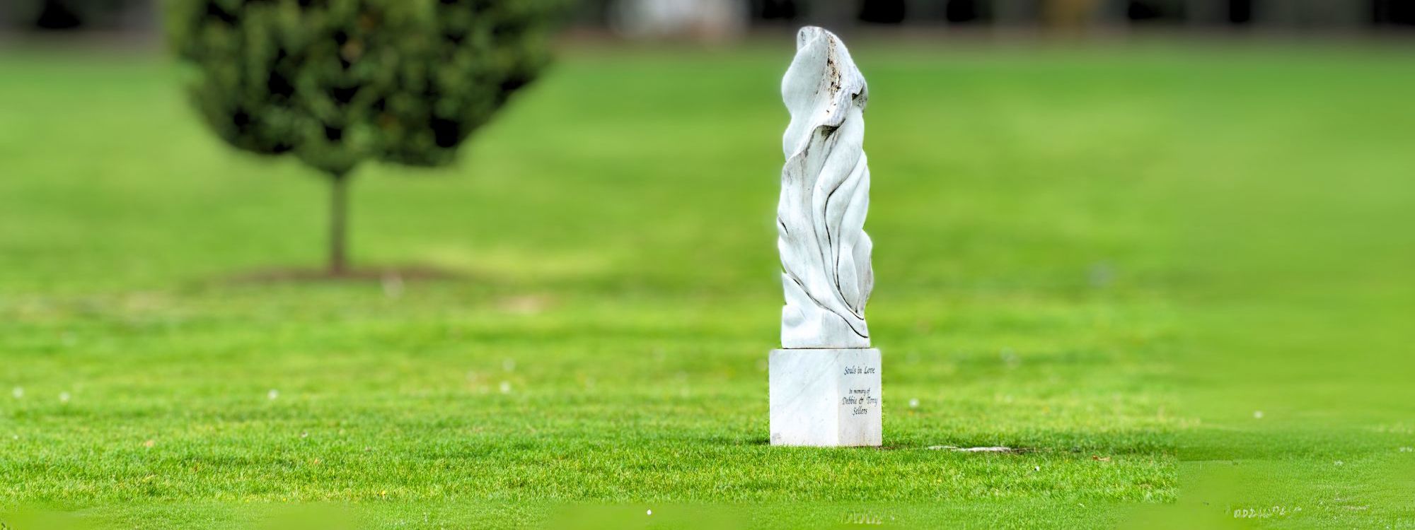 A serene park scene with a lush green lawn and a single, rounded tree on the left. On the right stands a smooth, white marble sculpture with abstract, flowing lines, possibly representing intertwined forms. The sculpture is inscribed with "Souls in Love. In memory of Debbie & Tony Salam." In the background, a row of tall, dark evergreen trees adds depth and contrast. The image exudes a peaceful and reflective atmosphere.