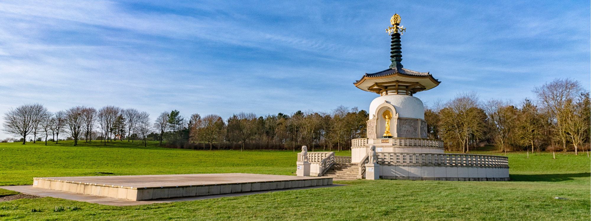 The image shows a white Buddhist stupa with a golden statue of Buddha placed in an alcove at the front. The stupa is situated in a grassy area with trees in the background under a blue sky with some clouds. The stupa features a traditional pagoda-style roof with a golden ornament at the top, and it is elevated on a platform with steps leading up to it. The overall design reflects elements of Japanese or East Asian architecture.