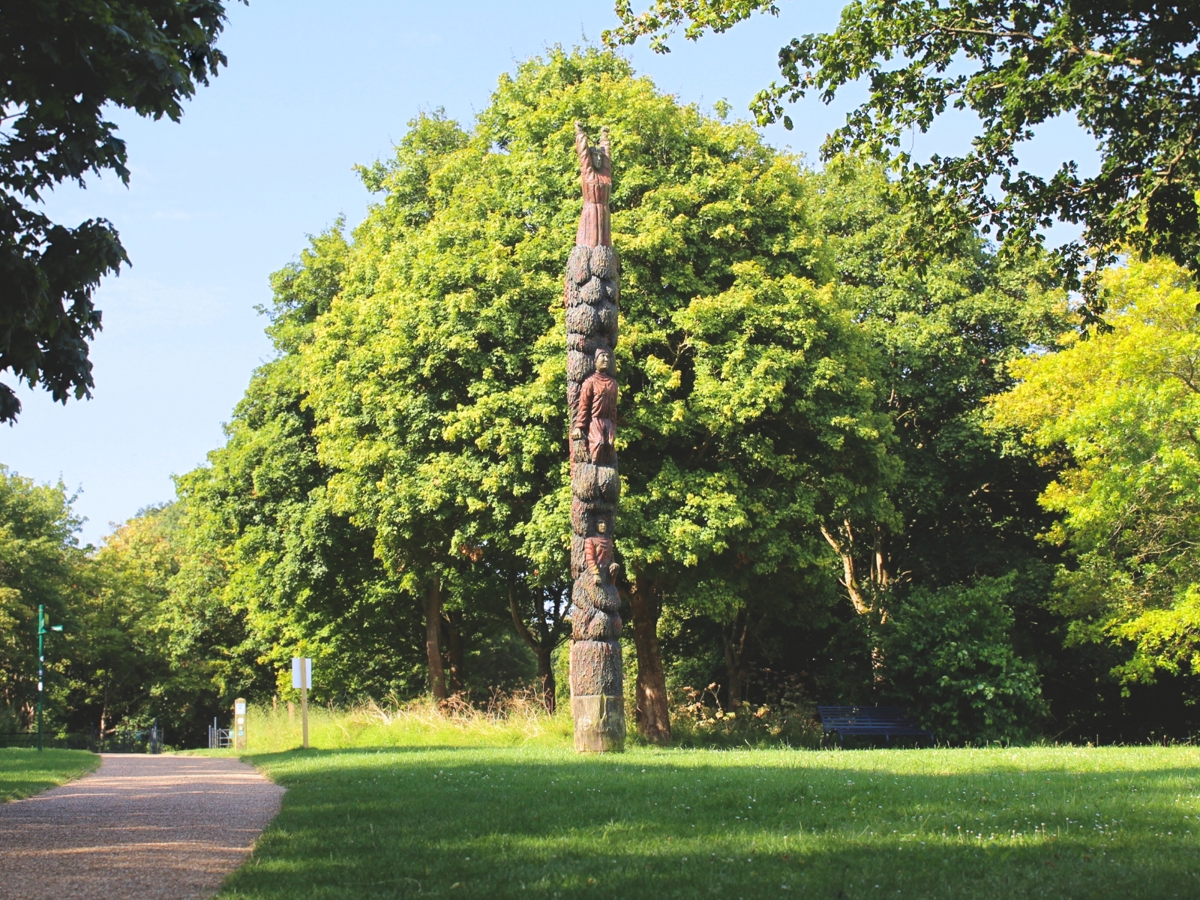 A tall wooden totem pole with carved and painted human figures stands upright in a grassy area surrounded by lush green trees. The totem pole is prominently positioned with a clear blue sky in the background