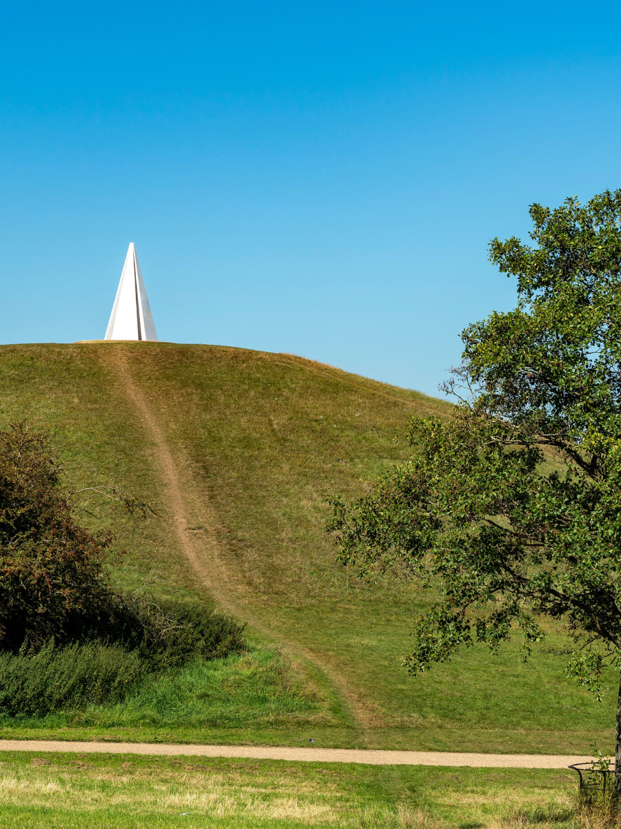 A large white pyramidal art sculpture on top of a big grassy hill. The sky is bright blue and sunny.