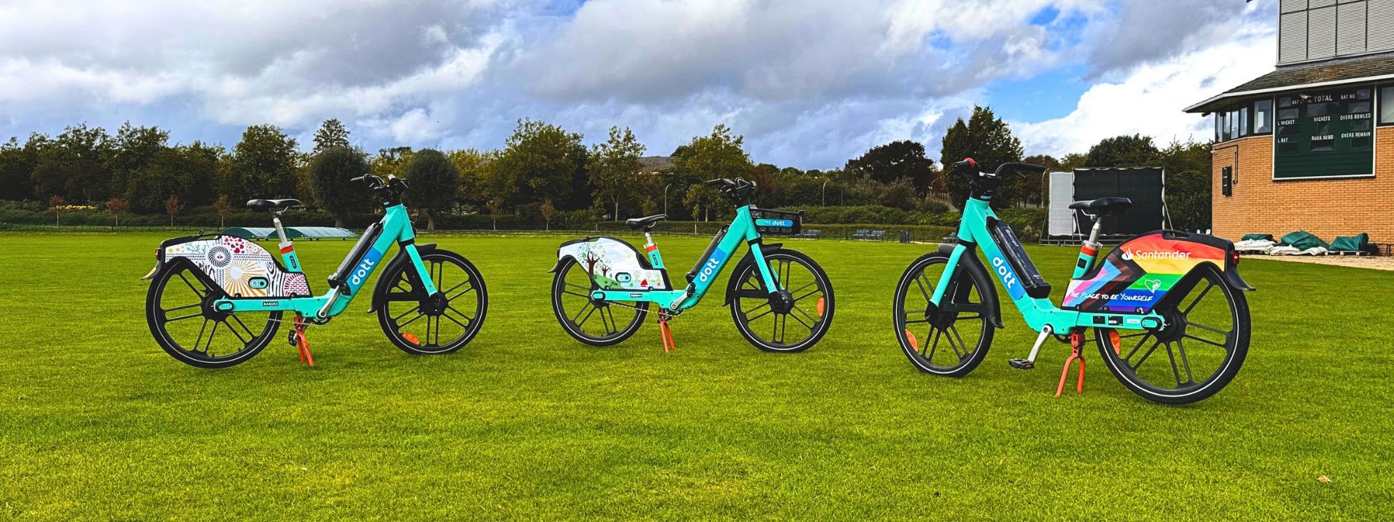 The image shows three bright blue rental bikes with large black wheels, standing in a row on a well-manicured green lawn under a partly cloudy sky. The bikes feature Santander branding, colorful designs on the rear fenders, and are equipped with red kickstands. A clock tower and scoreboard can be seen on the right side of the background, suggesting a sports field or park setting.