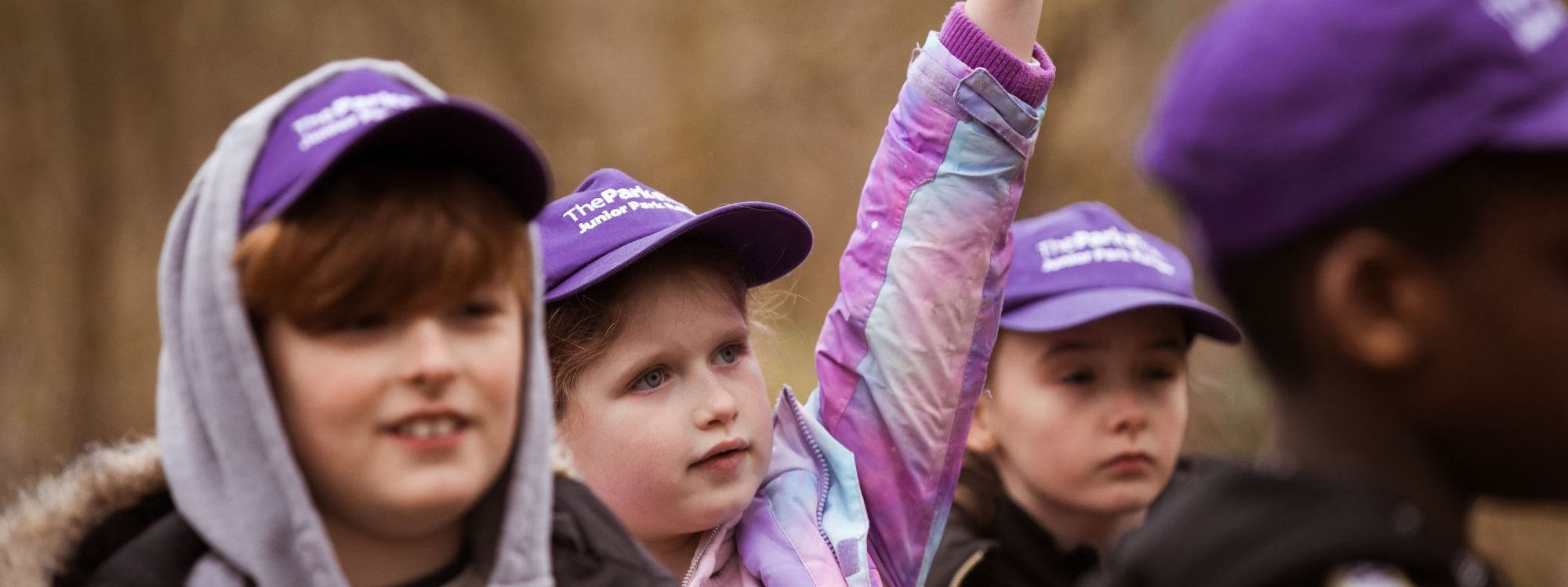 A group of children in purple hats is stood outside in a woodland. One child has her hand raised while another child, slightly out of focus, smiles into the distance.