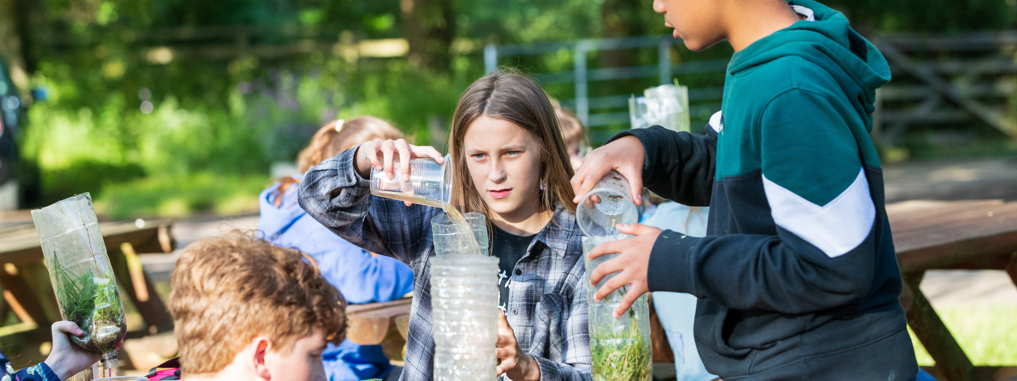 Three teenagers are sat at a bench outside making natural water filters. They are holding plastic cups of dirty water and pouring them through plastic bottles that contain leaves, grass and moss, among other things