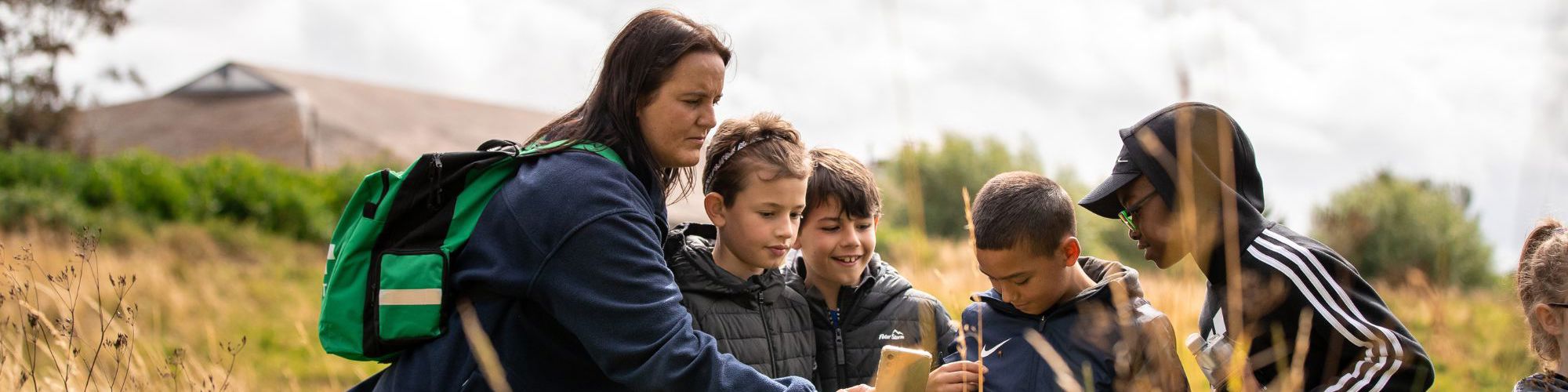 Teacher leading session with children in park. The female teacher is taking a photo on her phone, while standing in long brown grass. Four young boys are looking at the teacher's phone and smiling. 