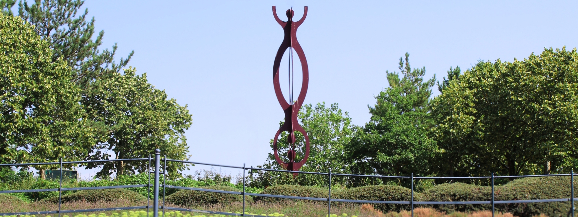 The image shows a tall, abstract metal sculpture with a deep reddish hue, prominently displayed in a landscaped area. The sculpture features two elongated, interconnected loops designed to look like people. The area surrounding the sculpture is fenced off with a black metal railing, and the ground is covered with low-lying greenery and small flowers. In the background, there are tall, leafy trees under a clear blue sky, creating a peaceful and well-maintained park-like setting.